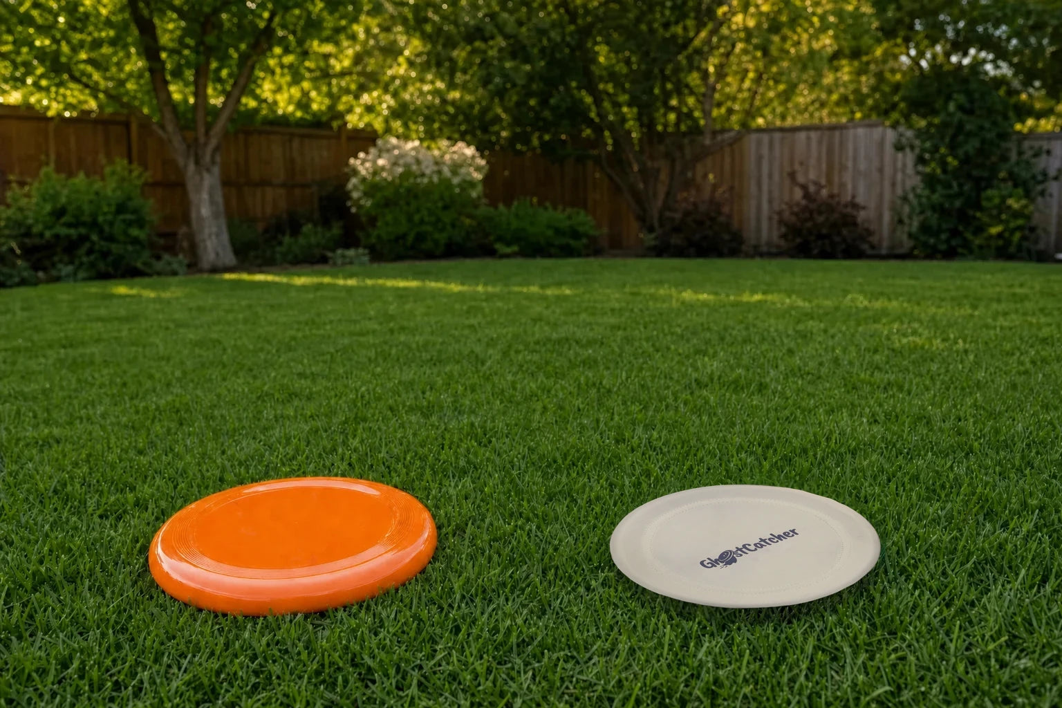 Two flying discs on a backyard lawn as a person sees them: a bright orange disc and an ivory GhostCatcher disc with its logo